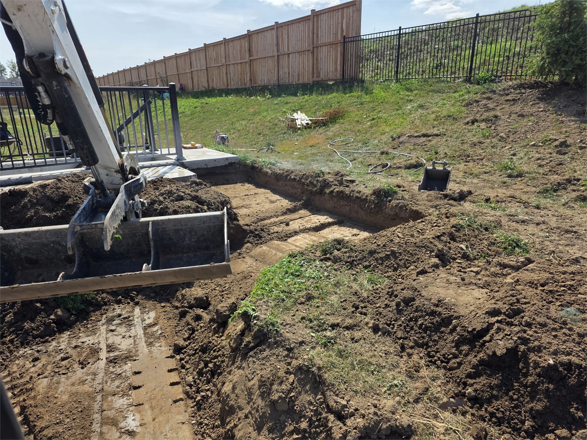 Excavator bucket over an excavated area beside a residential patio and fenced lawn.